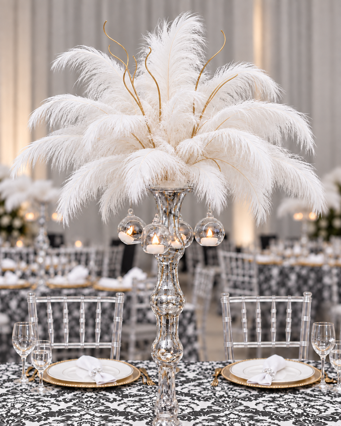 Full white ostrich feathers in a sculptural glass vase with hanging candle globes, charcoal damask linen, champagne-gold place settings and clear Chiavari chairs